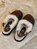 Zueco de cuero color suela con interior forrado en piel sintética blanca. Presenta una tira trasera ajustable con hebilla metálica y suela de goma negra con relieve.