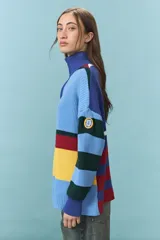 Sweater de tejido de punto grueso con diseño de bloques de color y rayas horizontales en tonos azul, rojo, amarillo y verde. Presenta cuello alto con cierre frontal parcial y mangas largas con puños ajustados.