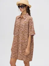 Camisa tipo vestido de corte oversize, confeccionada en viscosa. Presenta un diseño estampado con trazos abstractos en tonos verdes sobre una base naranja, cuello clásico, cierre frontal con botones y mangas cortas.