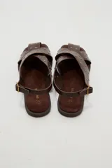 Sandalias planas de cuero sintético marrón, con tiras cruzadas en el empeine y tachas metálicas doradas distribuidas por toda la capellada. Cuenta con tira ajustable en el talón con hebilla.