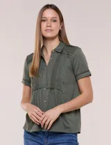 Camisa verde militar de manga corta, con textura flamé, cuello con solapa y cierre frontal de botones. Presenta alforzas verticales, detalles de puntilla calada y frunces bajo el busto que aportan volumen.