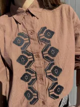 Camisa de algodón con corte clásico, cuello camisero y cierre frontal con botones. Presenta un diseño bordado en punto de cruz con motivos florales en color bordó sobre el frente. Cuenta con mangas largas con puños abotonados.
