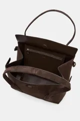Bolso tipo citybag confeccionado en serraje color marrón oscuro. Presenta una solapa frontal con cierre magnético, asas de hombro integradas y detalles de hebillas metálicas en los laterales para ajuste. Incluye bolsillo interior con cremallera.