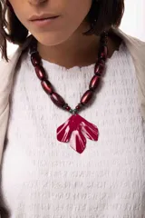 Collar corto con cuentas cilíndricas en tono bordó brillante y dije central en forma de hoja o flor de gran tamaño en color fucsia intenso. El diseño combina texturas y materiales artesanales.