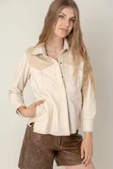 Camisa de cuero ovino color beige claro, estilo western, con canesú en los hombros en gamuza de tono similar. Presenta costuras contrastantes y cierre frontal con broches. Posee bolsillos laterales.