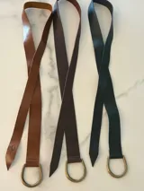 Cinto de cuero flexible y envolvente con diseño minimalista, que se anuda mediante una hebilla metálica en forma de D. El extremo presenta el logo de la marca grabado.