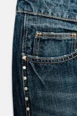 Jeans de tiro bajo y corte wide leg, confeccionados en denim azul con efecto lavado. Presentan detalle de tachas metálicas a lo largo de las costuras laterales y diseño clásico de cinco bolsillos.