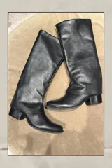 Bota de caña alta confeccionada en cuero vacuno negro, con diseño de solapa superpuesta en la parte superior de la caña y taco cuadrado bajo.