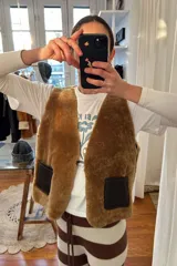 Chaleco reversible confeccionado en cuero genuino. Presenta un lado de piel de oveja gris con bolsillos de cuero negro y un lado de cuero liso gris. Diseño sin mangas con cierre frontal.