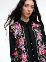 Vestido camisero largo de algodón negro con cuello camisero, cierre frontal de botones y mangas largas. Presenta un diseño con bordados florales coloridos a lo largo de toda la prenda.