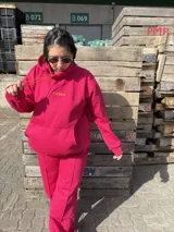 Canguro fucsia de tela deportiva con capucha, cordones ajustables en color naranja y logo bordado en el pecho.