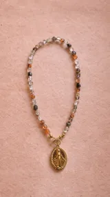 Collar compuesto por una cadena de piedras naturales facetadas en tonos translúcidos, beige y tierra, con un dije central de la Medalla Milagrosa en metal dorado.