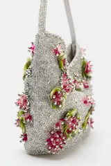 Bolso de hombro plateado cubierto de abalorios y cuentas, decorado con apliques florales tridimensionales en tonos verde y fucsia. Cuenta con un asa corta para el hombro, cierre de clip imantado y bolsillo interior con cremallera.