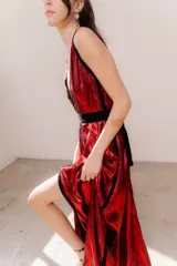 Vestido largo de seda color bronce con acabado brillante, escote profundo en V y espalda abierta. Presenta detalles en terciopelo negro que enmarcan el escote, recorren el frente y definen la cintura con una faja ajustable. Posee breteles anchos y una abertura frontal en la falda.