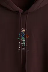 Canguro de corte holgado en color bordó, con capucha ajustable mediante cordones, hombros caídos y bolsillo frontal tipo canguro. Presenta un pequeño estampado central de un jarrón con flores coloridas.