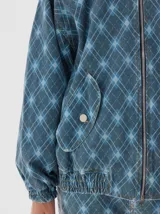 Campera bomber de denim con estampado de cuadros tipo argyle en tonos de azul. Presenta cierre metálico frontal, bolsillos delanteros con solapa y botón, y un ajuste oversized.