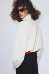 Chaqueta de punto color crudo con cuello alto, cierre frontal de cremallera metálica, mangas largas con puños elásticos y bajo ajustable con cordones.