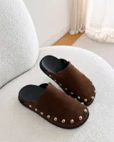 Zuecos de ante color beige con diseño destalonado, punta redonda y tachas metálicas plateadas alrededor de la suela.