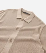 Camisa de punto de manga corta en color beige, con cuello inglés y cierre frontal de botones. Presenta una textura tejida y un corte regular que ofrece un ajuste cómodo y clásico.