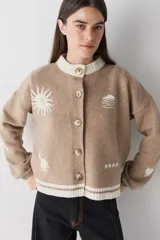 Cardigan de lana color crema con cuello redondo, cierre frontal de botones y mangas largas con puños ajustados. Presenta bordados decorativos en el frente que incluyen un sol, olas y estrellas, con detalles en azul en el cuello y el ruedo.