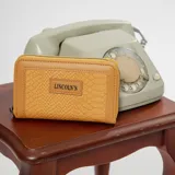 Billetera mediana con textura tipo croco, color mostaza, con cierre perimetral y placa metálica con logo de la marca en el frente.
