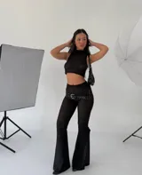 Pantalón de tiro medio con corte acampanado, confeccionado en tejido de encaje negro con textura. Presenta un cinturón ancho integrado con tachas metálicas y hebilla plateada.