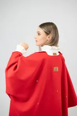 Capa de paño color rojo con corte amplio y caída natural. Presenta detalles de dijes metálicos en baño de oro distribuidos en la espalda y una etiqueta de cuero con el logo de la marca en la parte superior.