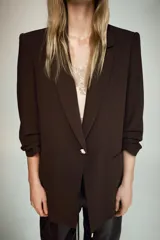 Blazer negro de cuello solapa con hombreras, manga por debajo del codo con drapeado y falsos bolsillos de vivo en el delantero. Cuenta con forro interior a tono y cierre frontal con un botón.