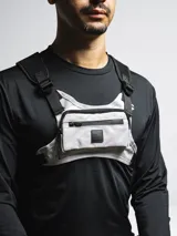 Bolso de pecho táctico color gris claro con detalles en negro, con correas ajustables y hombreras de neopreno. Cuenta con tres compartimentos: uno trasero de red y dos frontales con cierre.