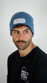 Gorro tipo beanie de punto acanalado color celeste, con parche rectangular frontal que incluye el logo y nombre de la marca Duxon.