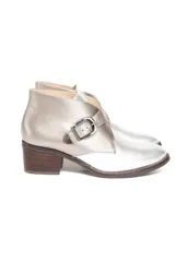 Bota corta de cuero color plateado metalizado, con taco cuadrado de madera, punta redonda y diseño cruzado con hebilla lateral plateada.