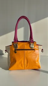 Cartera de cuero estilo tote de la marca Carolina De Cunto, confeccionada con una combinación de cueros en tonos azul, fucsia, beige y dorado. Cuenta con asas de mano fucsias, detalles de pelo sintético en el frente, bolsillos externos con cierre y herrajes metálicos.