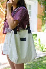 Bolso tipo tote color arena con asas de cuero sintético negro y parche con logo en el frente.