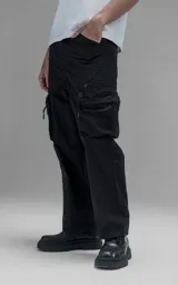 Pantalón cargo oversize de corte relajado, confeccionado en textil técnico resistente de color negro. Presenta bolsillos laterales cargo de gran tamaño con cierres metálicos, bolsillos laterales superiores y bolsillos traseros con broches a presión. Incluye cinturón integrado de cinta de seguridad con hebilla de encastre técnica.