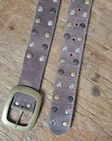 Cinto de cuero marrón con hebilla metálica de estilo envejecido y detalle de tachas metálicas en diferentes tonos en el extremo.