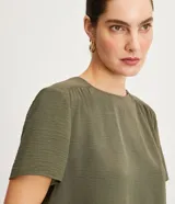 Blusa de poliéster color verde oliva, con cuello redondo, manga corta y detalle de pliegues fruncidos en la costura de los hombros.