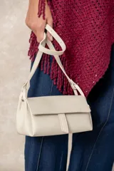Cartera pequeña de hombro en color beige, confeccionada en cuero sintético con textura granulada. Presenta un diseño rectangular con solapa frontal y cierre magnético, e incluye una correa ajustable.