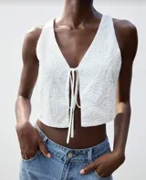 Chaleco crop top blanco con bordado floral y lazos frontales.