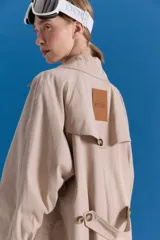 Trench midi de color camel, confeccionado en una mezcla de algodón y nylon con propiedades repelentes al agua. Presenta un diseño clásico con cuello camisero, cierre frontal de botones, bolsillos laterales y tajos en los costados.