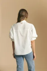 Camisa blanca de manga corta con volumen en los hombros, cuello camisero y cierre frontal con botones dorados.