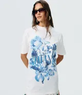 Remera de algodón blanca de corte largo, manga corta y cuello redondo. Presenta un gran estampado floral en tonos azules con la inscripción "Botanist" en el frente.