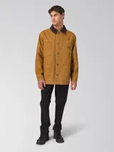 Chaqueta estilo chore de lona color marrón con cuello camisero de pana marrón oscuro, cierre frontal con botones y cuatro bolsillos de parche delanteros con pespuntes contrastantes blancos.