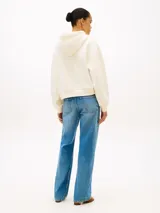 Buzo con capucha Tommy Hilfiger de color blanco, confeccionado en felpa de puro algodón. Presenta un corte amplio con hombros caídos, puños y bajo de punto canalé, y cordón ajustable en la capucha. En el pecho destaca un logotipo texturizado en tono rojo, complementado con un pequeño logo bordado en el puño.