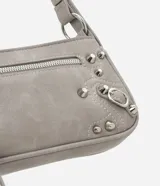 Cartera tipo baguette de PU color beige claro, con acabado liso y detalles de apliques metálicos tipo tachas y argollas en color gunmetal. Presenta un bolsillo frontal con cierre y un tirador de cuero sintético colgante. Incluye asa corta ajustable con hebilla.