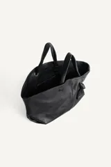 Bolso tipo shopper de cuero negro con textura granulada. Presenta doble asa de mano, cierre magnético y un detalle decorativo de nudo con argolla metálica en el frente.