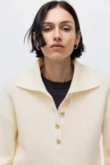 Buzo de punto acanalado color beige, con cuello polo y tapeta frontal con botones metálicos decorativos. Presenta hombros caídos, mangas largas con puños ajustados y diseño de corte recto.