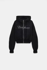 Sudadera negra con capucha, cierre frontal de cremallera metálica y mangas largas con puños. Presenta un diseño corto con bolsillo tipo canguro y detalle de texto 'California' aplicado con brillos en el frente.