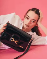 Cartera negra de cuero charolado con solapa y cierre de broche. Presenta un detalle metálico dorado en forma de freno de caballo en la parte frontal y una correa ajustable.