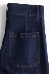 Jean de tiro alto en denim azul oscuro con pierna amplia y corte tobillero. Presenta un diseño de cinco bolsillos, cierre frontal con botón y costuras decorativas verticales en el frente que simulan un pliegue.