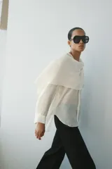 Camisa de hilatura con mezcla de lyocell color blanco, con cuello fluido con pañuelo incorporado, manga larga con puño y bajo redondeado asimétrico. Cierre frontal con botones ocultos.